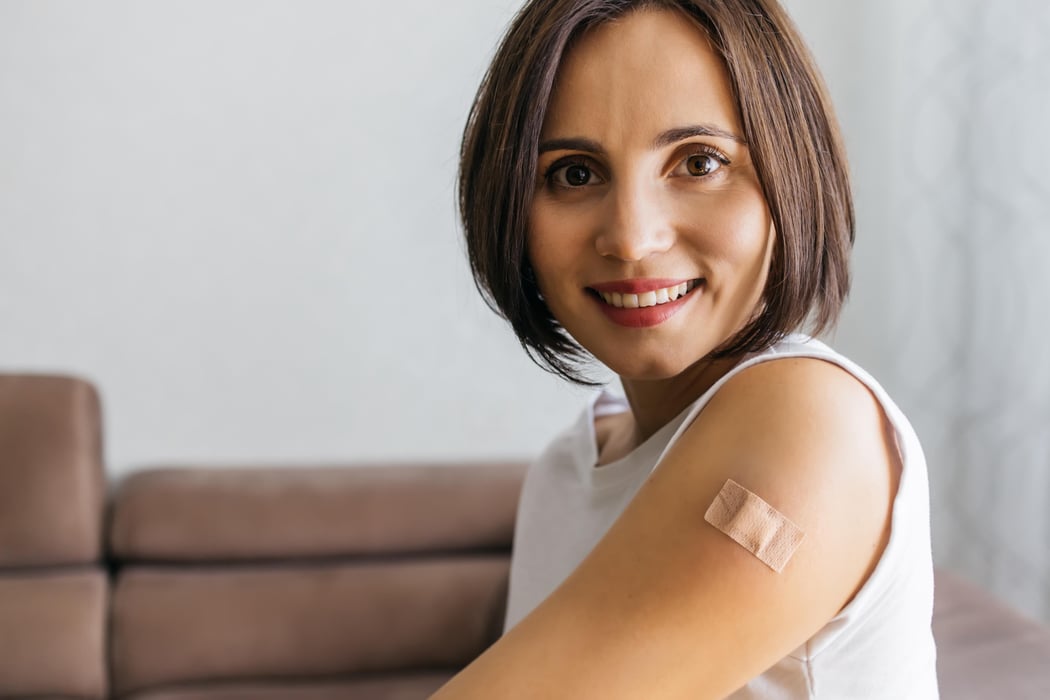 Portrait of smiling woman after getting a vaccine. Female holding down her white shirt sleeve and showing her arm with bandage after receiving vaccination. Concept of recommended inoculation