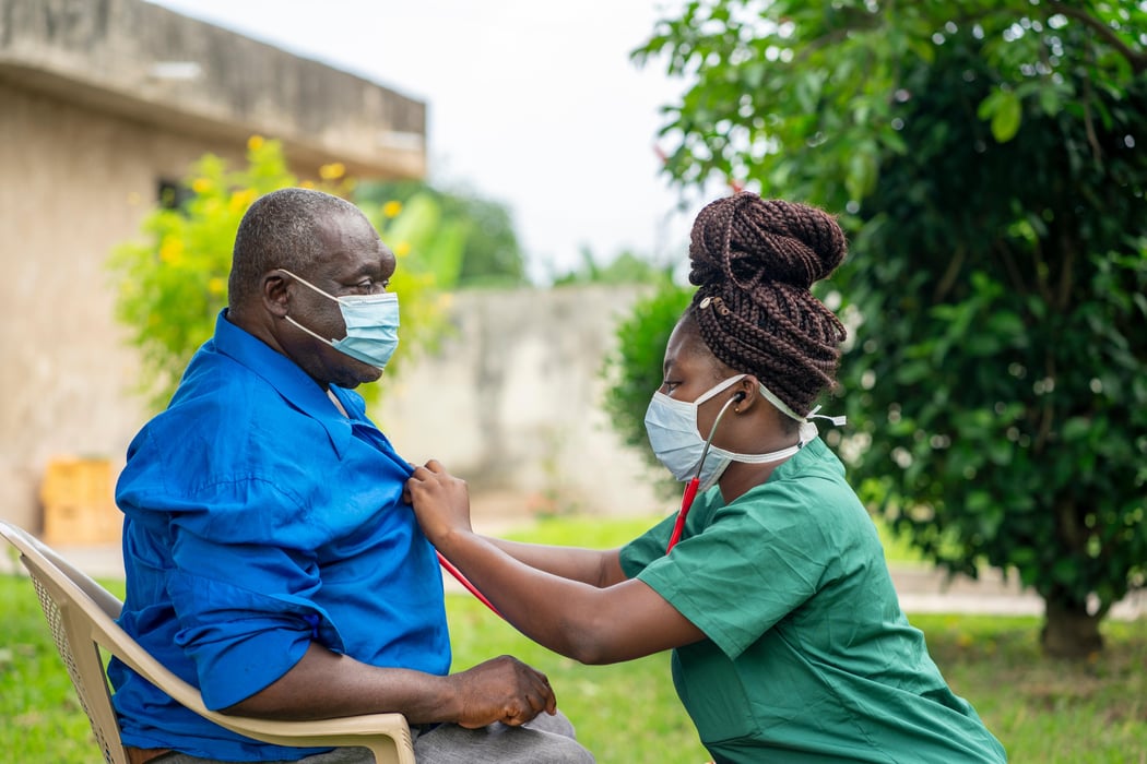 image of an aged African man being checking up by a health worker-young black nurse wearing a face mask taking care of an old man- health worker at home service