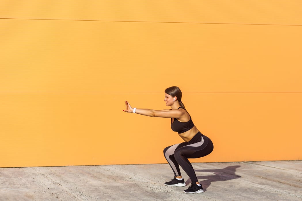 Brunette young woman doing a squat outside in front of an orange wall
