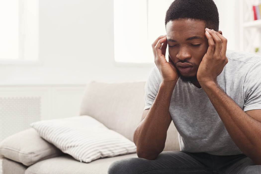 black young man sitting on white couch with head in hands with anxiety