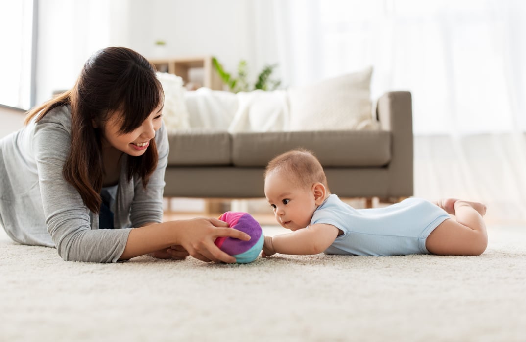 mom lying on her stomach on white rug with baby on their stomach playing with a soft rainbow ball